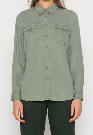 Frau trägt ein olivgrünes, langärmeliges Hemd mit Knöpfen und zwei Brusttaschen sowie schwarze Hosen, steht vor einem einfarbigen Hintergrund.