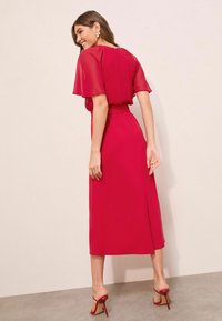 Femme souriante, portant une robe midi rouge à manches courtes transparentes et des sandales à talons rouges, se tenant devant un mur clair.