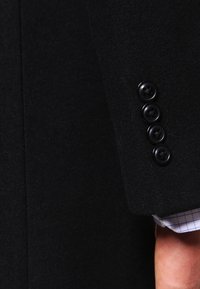 Zwarte wollen blazer met een keurig getailleerde mouw met vier zwarte knopen en een geruit shirtmanchet dat eronderuit komt. Glad, gestructureerd materiaal.