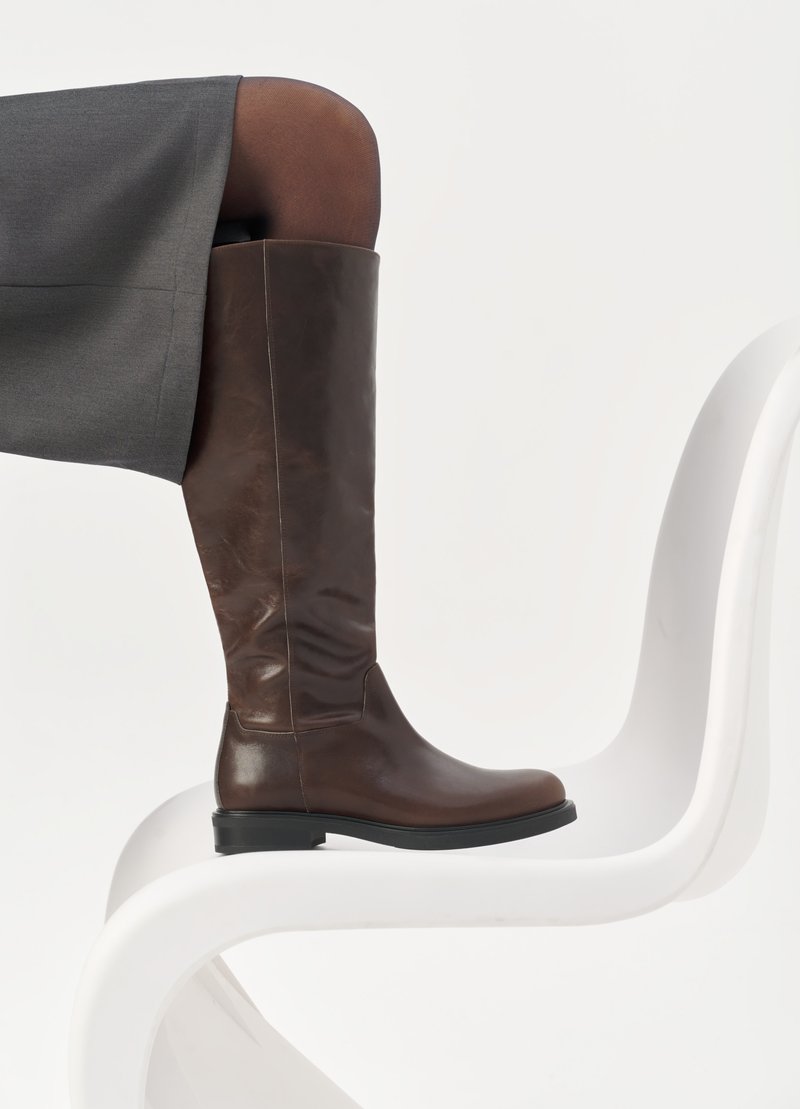 Botte en cuir marron à hauteur de genou avec un bout arrondi, texture lisse et détails de couture minimalistes, placée sur un fond clair sur une chaise blanche.