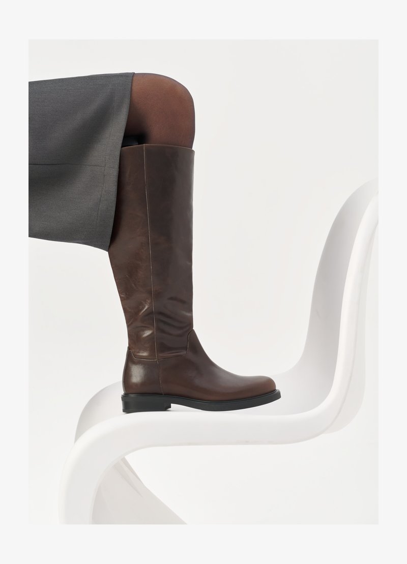 Stivale in pelle marrone alto fino al ginocchio con punta arrotondata, superficie liscia e dettagli di cucitura minimi, posato su uno sfondo chiaro su una sedia bianca.