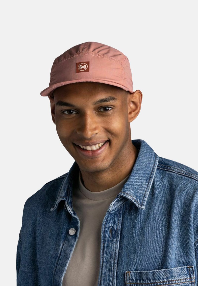 Gorra rosa con visera curva y un parche con logo, llevada por una persona con chaqueta de mezclilla. Tela suave, diseño flexible, aspecto casual.
