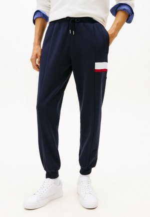 Marinefarbene Jogginghosen mit Seitentaschen, Kordelzugbund und weißen sowie roten Streifenakzenten. Kombiniert mit weißen Sneakers. Weicher Stoff, schmale Passform.