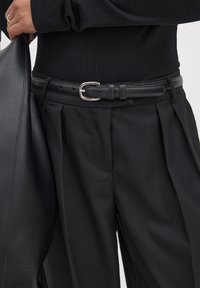 Black ribbed sweater paired with high-waisted black trousers, featuring pleats and a black leather belt with a silver buckle. A black bag is visible.