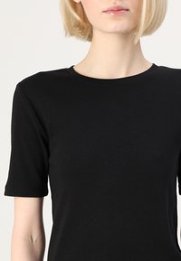 Černé bavlněné tričko s kulatým výstřihem a krátkými rukávy, které má hladkou texturu a jednoduchý design, zobrazené od hrudníku nahoru.
