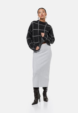 Femme portant un pull à carreaux noir et gris, une jupe crayon gris clair, des bottes noires à talons hauts et des boucles d'oreilles créoles, debout sur un fond blanc.