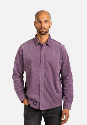 Kahlköpfiger Mann mit Ohrringen, der ein violettes Hemd mit Knöpfen und schwarze Jeans trägt, steht mit einer Hand in der Tasche vor einem weißen Hintergrund.