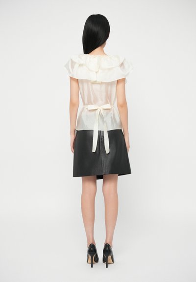 Sieviete ar garu melnu matu, valkājot caurspīdīgu balto blūzi ar volāniem, piesiets pie jostas, melnu ādas svārku un melniem augstiem papēžiem, stāv uz balta fona.