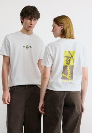 Giovane uomo di fronte e giovane donna di lato che indossano t-shirt bianche con la scritta "New York City" e pantaloni marroni su sfondo bianco.