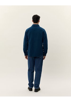 Homme debout, de dos, portant une veste polaire bleue, un jean bleu et des chaussures noires sur un fond clair.