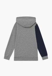 Sudadera con capucha y mangas largas, cuerpo y manga izquierda de color gris claro, manga derecha azul marino oscuro, puños y dobladillo acanalados, vista trasera.