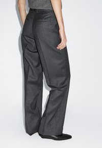 Grau angepasste Hose mit weitem Bein. Merkmale sind zwei Gesäßtaschen und eine glatte Textur, kombiniert mit schwarzem Schuhwerk.