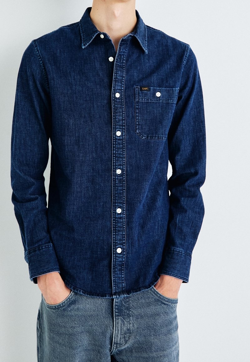 Homme portant une chemise en jean bleu foncé à boutons avec poche avant, associée à un jean bleu clair, mains dans les poches, sur fond uni.