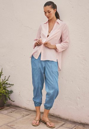 Frau mit langen dunklen Haaren, die ein pinkfarbenes Hemd, hellblaue kurze Hose und beige Sandalen trägt, steht vor einer hellen, strukturierten Wand.