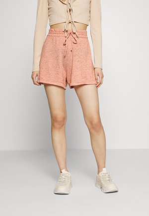 Personne portant un short taille haute rose pâle avec cordon, un top beige court à manches longues et des baskets épaisses beige clair, debout sur un sol gris.