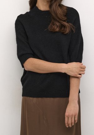 Vrouw met zwart kort mouwensweater en bruine rok, met lang bruin haar, haar armen voorzichtig over haar lichaam gekruist.