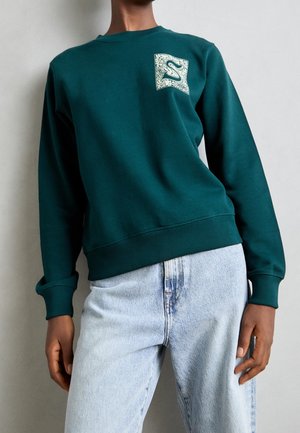 Person trägt ein türkisfarbenes Sweatshirt mit weißem grafischem Aufnäher auf der Brust und hellblaue, hoch taillierte Jeans, steht vor einer einfachen Wand.