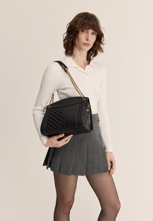 Borsa a tracolla in pelle nera trapuntata con una forma strutturata, tracolla in catena dorata, e una gonna grigia plissè indossata da un modello su uno sfondo neutro.