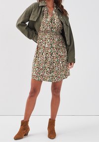 Femme portant une courte robe à fleurs avec une veste verte courte et des bottines en daim marron, se tenant devant un fond clair.