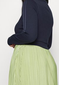 Mörkblå ribbad långärmad topp med hög krage, i kombination med en ljusgrön veckad kjol med vertikala linjer och en flödande textur.