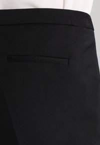 Black tailored pants feature a back welt pocket and a waistband with textured side insert, worn with a tucked-in white shirt.