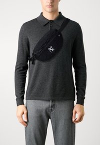 Svart fleece crossbody-väska med dragkedja, texturerad yta och broderad logotyp, bärs över en mörkgrå långärmad tröja.