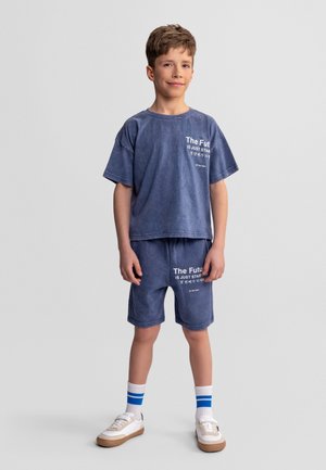 Chlapec stojí, oblečený v modrých zodpovedajúcich šortkách a tričku s bielym textom, biele ponožky s modrými pásikmi a biele tenisky.