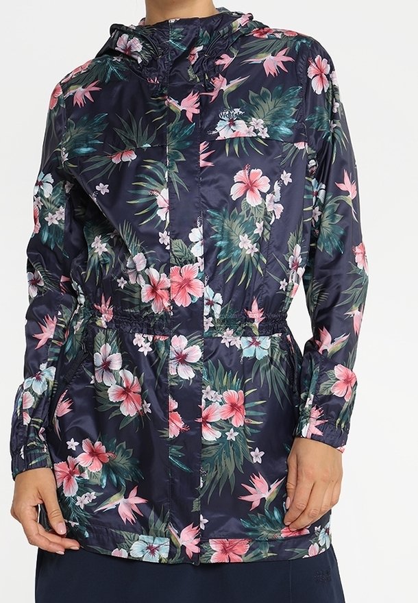 Femme portant une veste de pluie fleurie bleu marine avec des fleurs roses et blanches, manches longues, capuche et taille élastique, debout les mains près des poches.
