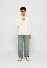 Hvit teksturert genser med høy krage, paret med lys blå løse denimjeans og kremfargede sko, stående foran en nøytral bakgrunn.