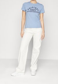 T-shirt di cotone azzurro chiaro con la scritta "ORIGINAL GANT SPORTSWEAR" in lettere blu navy, abbinata a pantaloni bianchi a gamba larga e sneaker bianche.