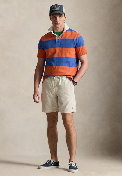 Νέος άνδρας που φοράει ριγέ πουκάμισο ράγκμπι σε μπλε και πορτοκαλί χρώμα, μπεζ σορτς, ναυτικό μπλε αθλητικά παπούτσια και καπέλο Polo Team σε ναυτικό μπλε, στέκεται μπροστά σε ένα απλό φόντο.