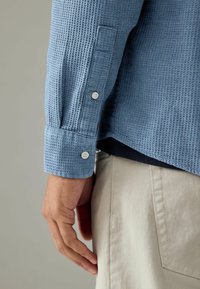 Cămașă lungă cu mâneci, textura albastru deschis, cu manșete cu nasture, asociată cu pantaloni bej deschis, având un tiv drept și un model subtil.