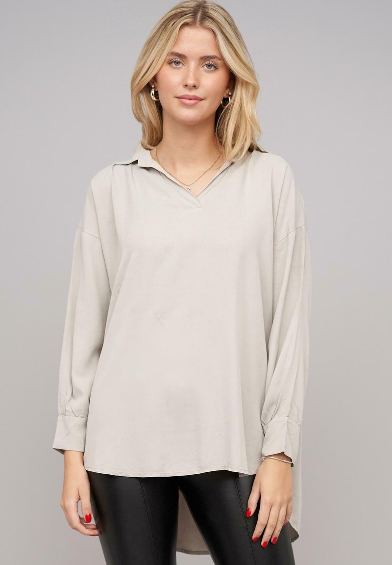 Blusa grigia chiaro a maniche lunghe con scollatura a V, vestibilità casual, texture morbida e orlo leggermente curvato; abbinata a pantaloni neri aderenti.