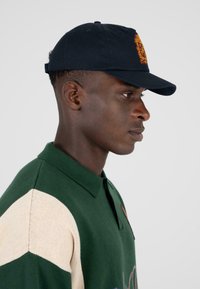 Marineblauwe baseballcap met geborduurd ontwerp; groen en crèmekleurig gebreid poloshirt met een kraag en gestructureerde stof; zijaanzicht.