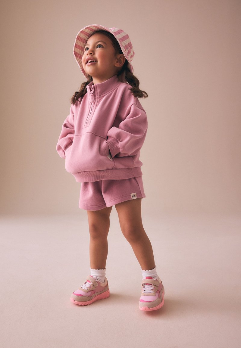 Niña joven con sudadera rosa, pantalones cortos a juego, gorra a rayas y zapatillas rosas que está de pie mirando hacia arriba y sonriendo con las manos en los bolsillos.