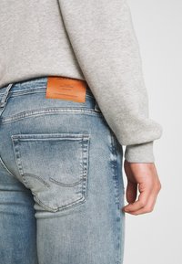 Ljust blå denimjeans med en blekt finish, bakfickor och en orange läderlogotyp synlig ovanför midjebandet, kombinera med en grå sweatshirt.
