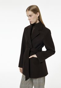 Brązowy płaszcz z paskiem, z wyciętym kołnierzem, bocznymi kieszeniami i długimi rękawami, w połączeniu z szarymi spodniami w kratę. Gładka faktura.