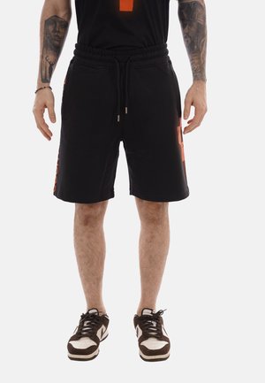 Mand iført sorte shorts med snøre og orange tekst på siden, sorte og hvide sneakers og synlige tatoveringer på armene, stående på en hvid baggrund.