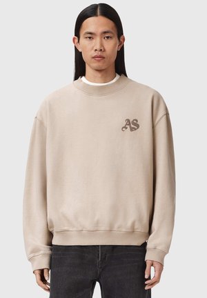 Jeune personne aux longs cheveux noirs portant un sweat-shirt beige oversize avec les initiales "AS" sur la poitrine et un pantalon noir.