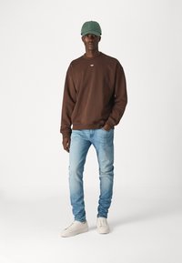 Felpa marrone con collo a giro, jeans azzurri e sneakers bianche. Un cappello verde completa l'outfit. Design semplice e casual.