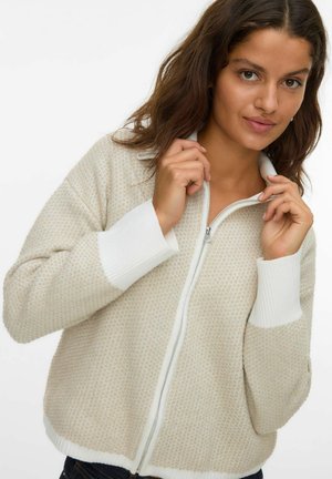 Kvinde med brunt hår iført en beige zip-up sweater med hvide manchetter, som holder og justerer kraven mod en ensfarvet hvid baggrund.