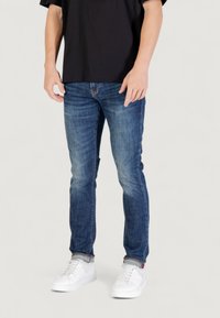Hombre vistiendo vaqueros ajustados azul, camiseta negra y zapatillas blancas, de pie contra un fondo claro liso.