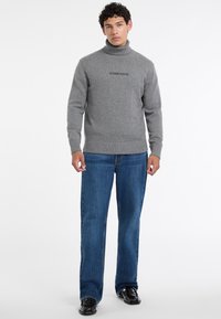 Maglione grigio a collo alto in una texture morbida con polsini e orlo a costine. Presenta il logo "GUESS JEANS" in nero. Abbinato a jeans blu a gamba dritta.
