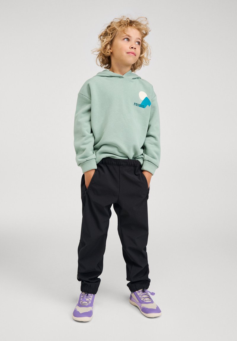 Παιδί με σγουρά ξανθά μαλλιά, φοράει φουτερ μπουφάν μέντας, μαύρο παντελόνι φόρμας και μοβ αθλητικά παπούτσια, στέκεται με τα χέρια στις τσέπες.