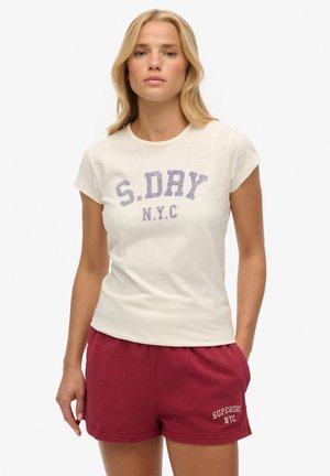 Kvinde med langt blond hår iført en hvid S. DRY NYC T-shirt og røde shorts med teksten Superdry NYC, står med hænderne i lommerne.