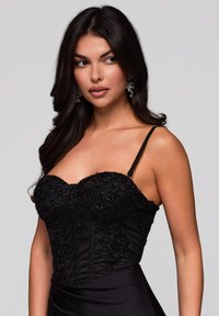 Kvinna med långt mörkt hår bär en svart dekorerad strapless topp och iögonfallande örhängen, tittar något åt vänster mot en enkel bakgrund.