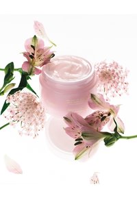 Roze crème in een doorschijnende pot, omringd door delicate roze bloemen. De pot heeft minimale labeling en een gladde, glanzende deksel.