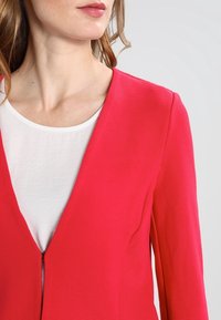 Světlý růžový blazer s ribovanou strukturou, s hlubokým výstřihem do V a zipovým zapínáním vpředu. Nositý přes bílý top.