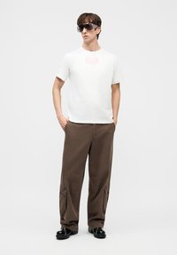 Homme portant un t-shirt blanc à manches courtes, un pantalon cargo marron à jambes larges, des chaussures noires, et de grandes lunettes de soleil foncées, debout les mains dans les poches.