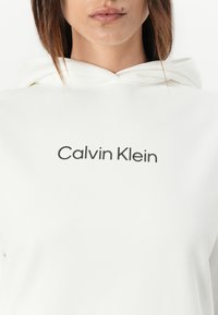 Felpa bianca realizzata in morbido tessuto, con una stampa frontale del logo "Calvin Klein" in un carattere elegante e moderno. Design semplice con cappuccio regolabile.